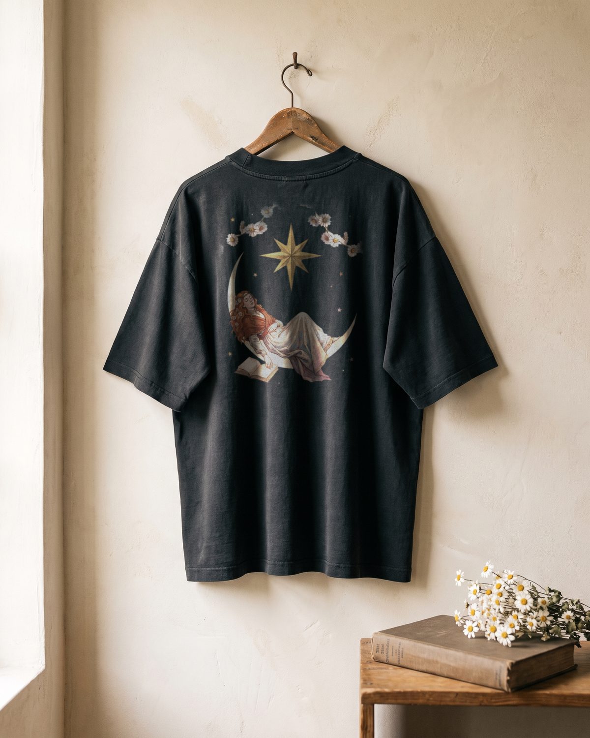 Mockup of the back of a faded black oversized tee hanging on a wall. The print shows a copper-haired woman asleep on a crescent moon with an open book, a gold eight-point star above her and a garland of daisies.