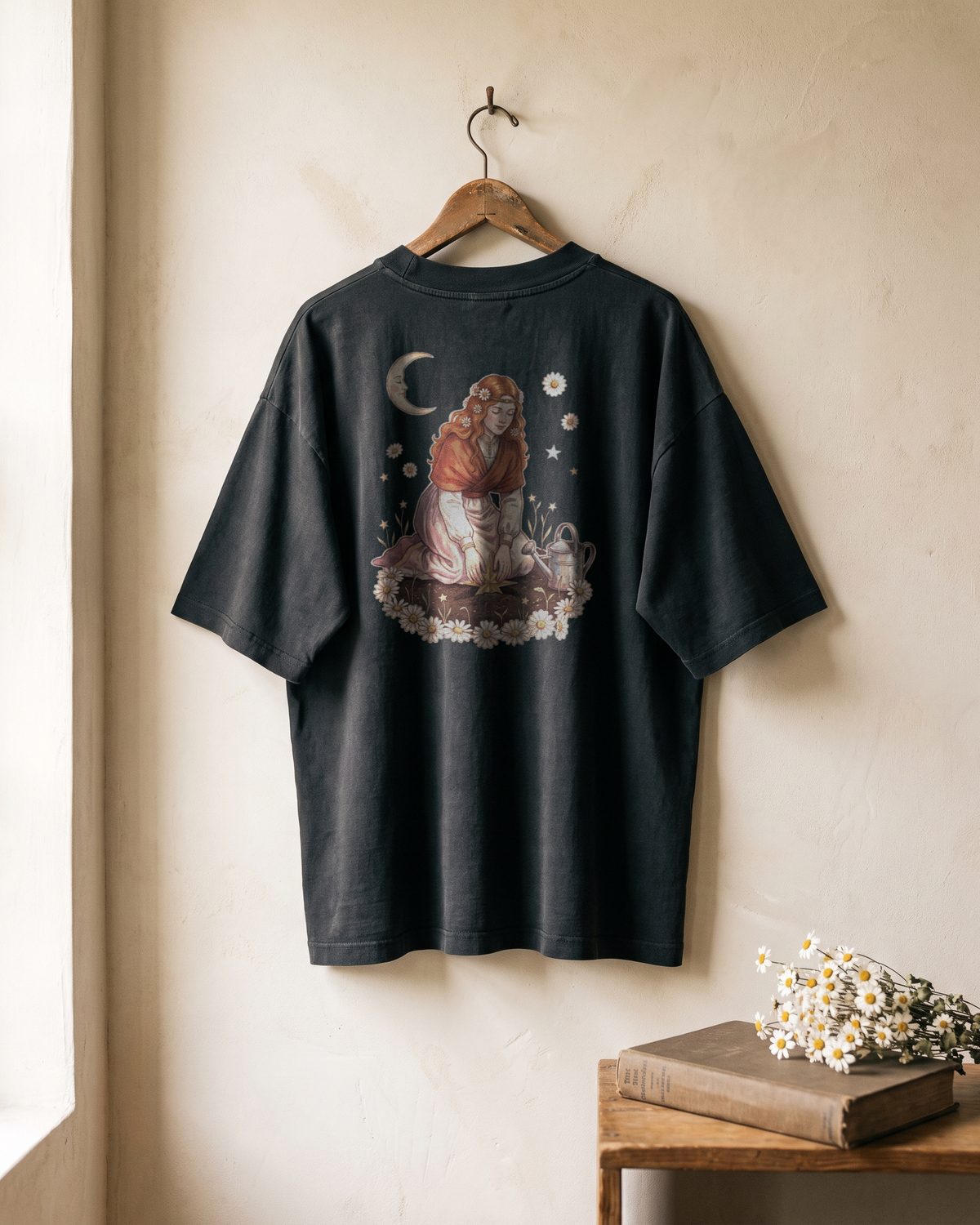 Mockup of the back of a faded black oversized tee hanging on a wall. The print shows the copper-haired woman kneeling among daisies, planting a small gold star in the soil like a seed.
